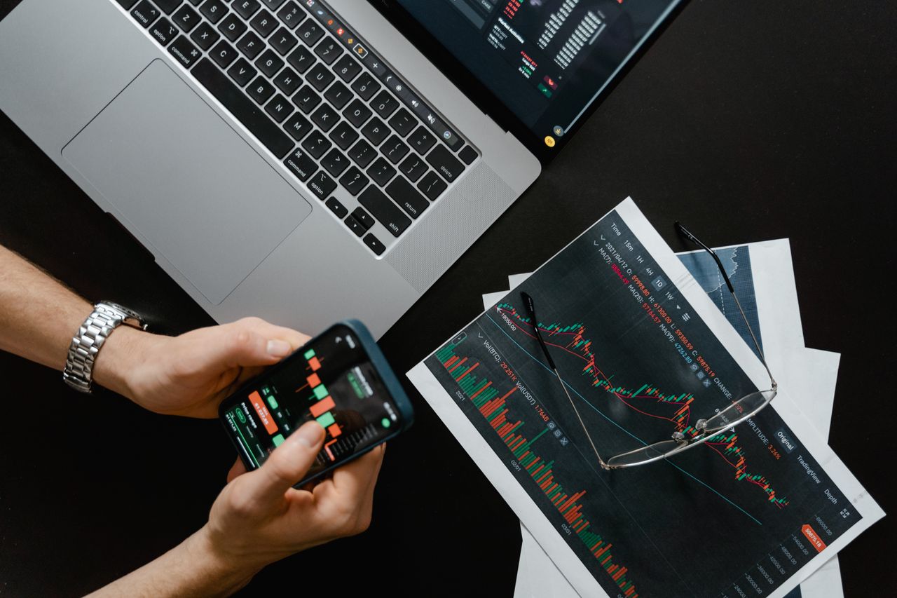 contact-img Des mains utilisent un smartphone à côté d’un ordinateur portable avec des graphiques boursiers, illustrant le trading numérique.