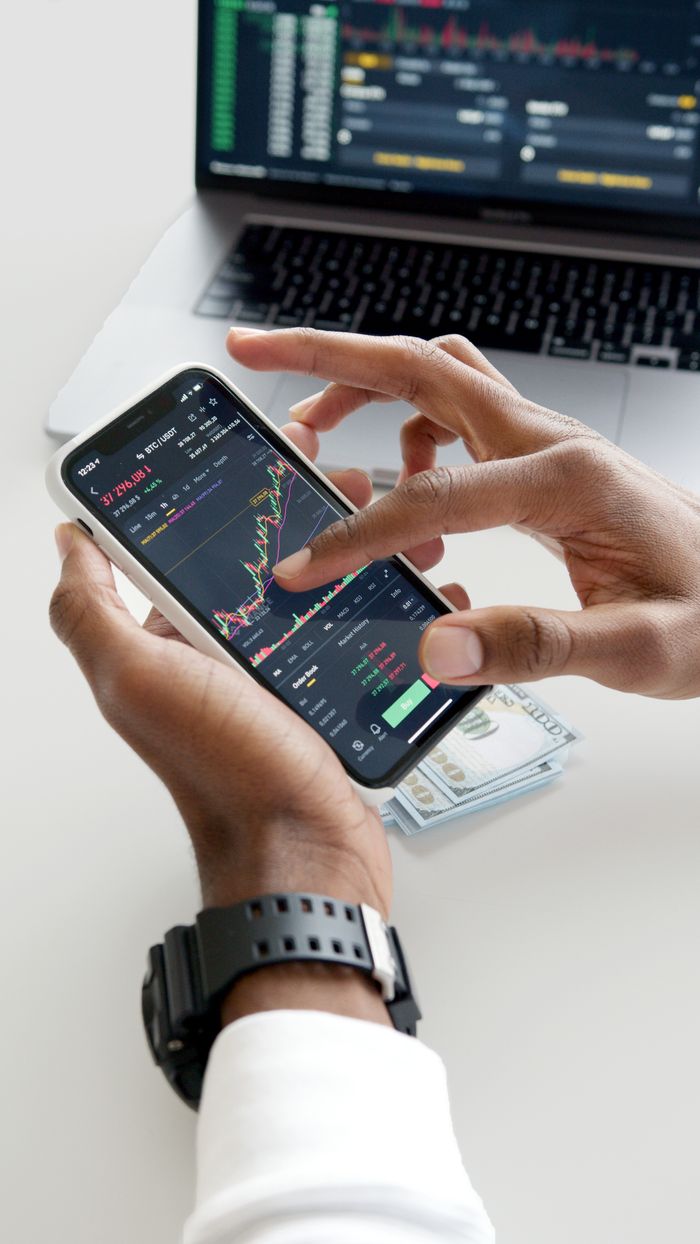 about-01 Des mains utilisant une application de trading sur smartphone avec des graphiques et des billets de dollar en arrière-plan.